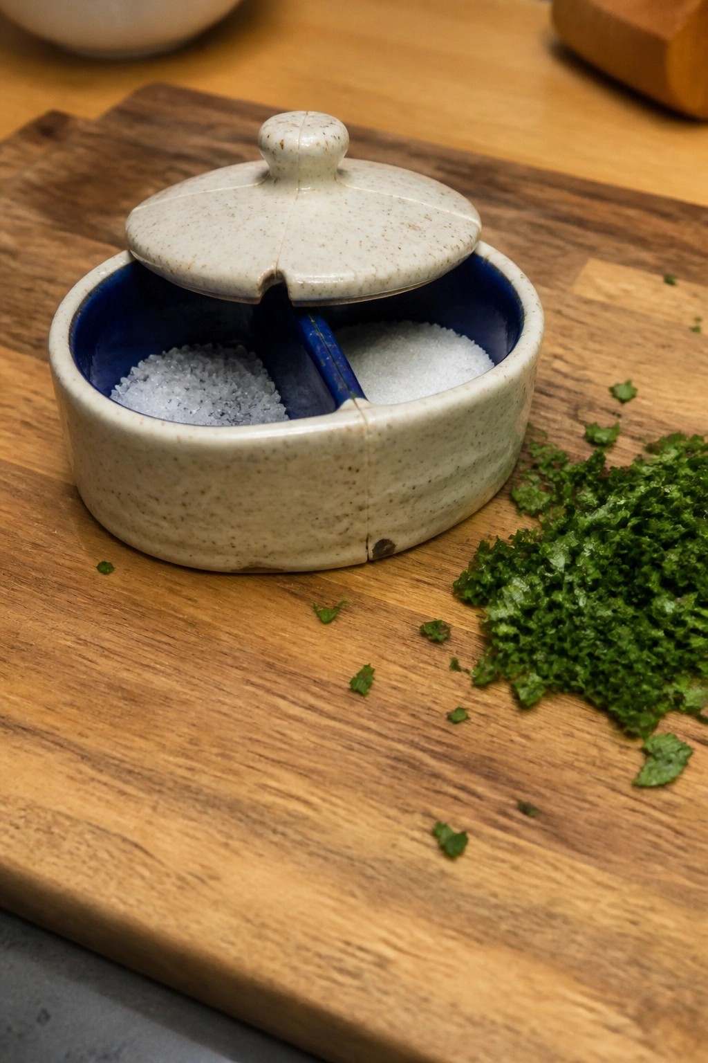 Small handmade ceramic container with two compartments and a lid, filled with white salts on a wooden surface beside chopped green herbs.