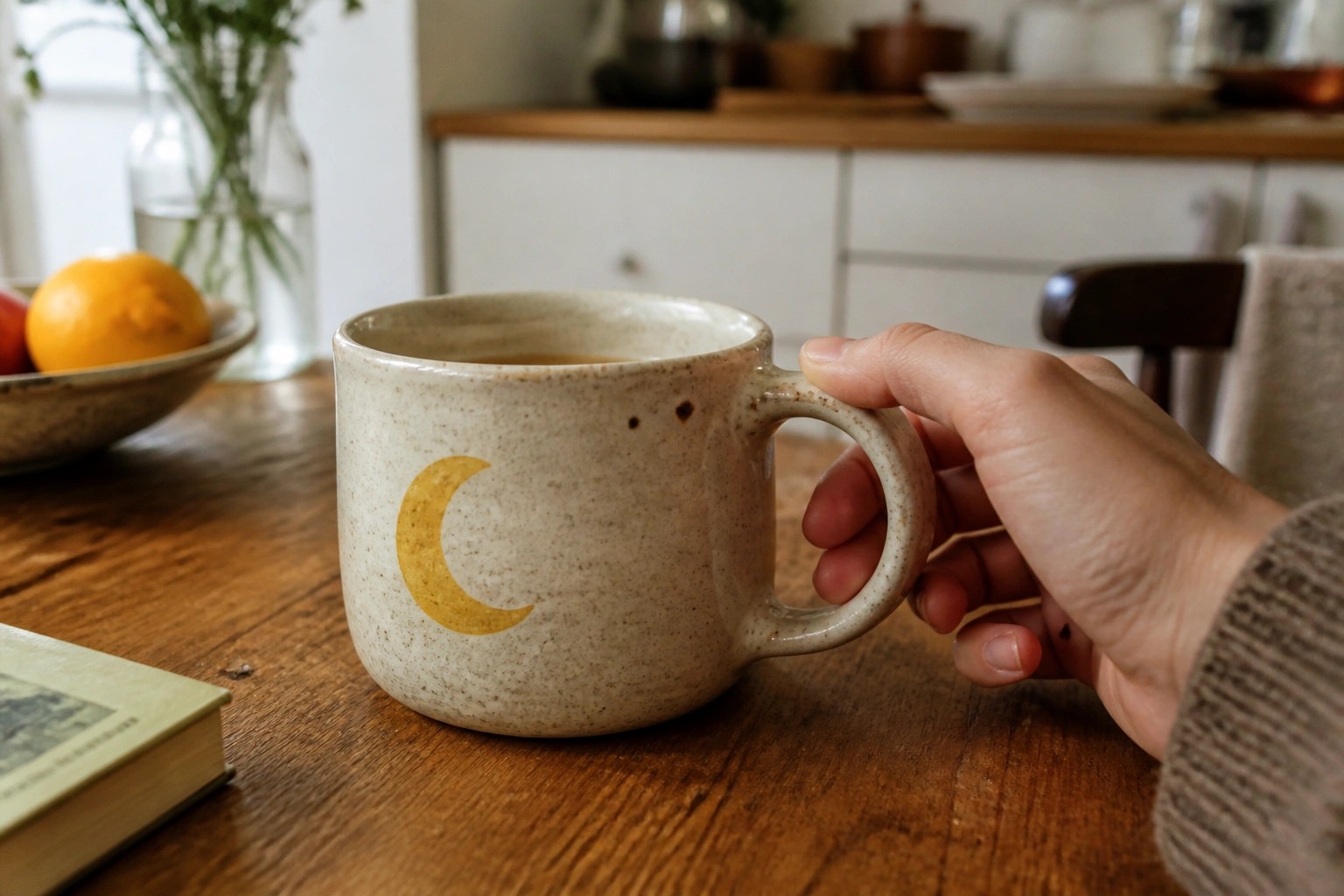 Hand in beige knit holding speckled mug with golden crescent moon on wooden table