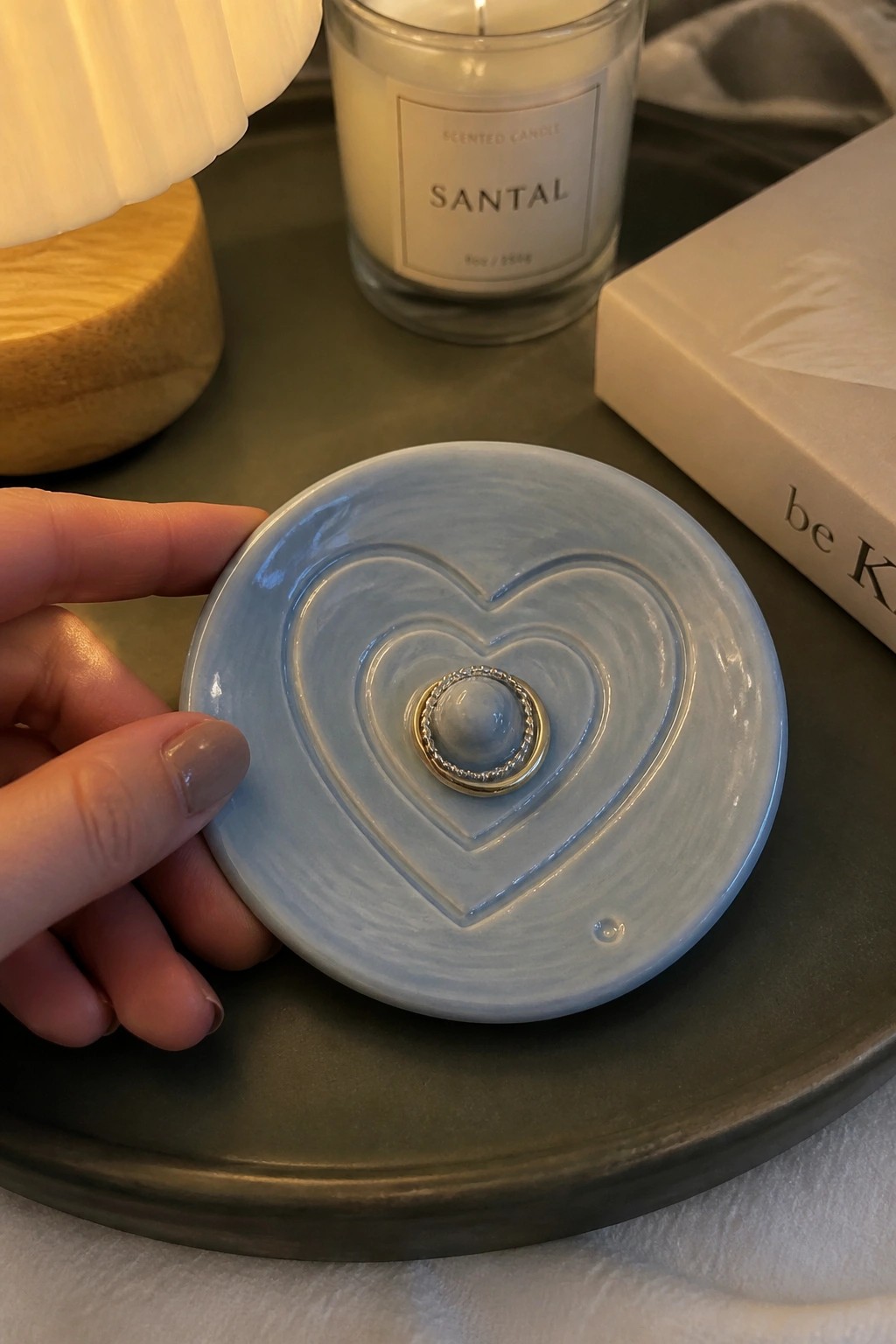 A hand holding a small light blue ceramic dish with a carved heart in the center and a ring resting inside the heart.