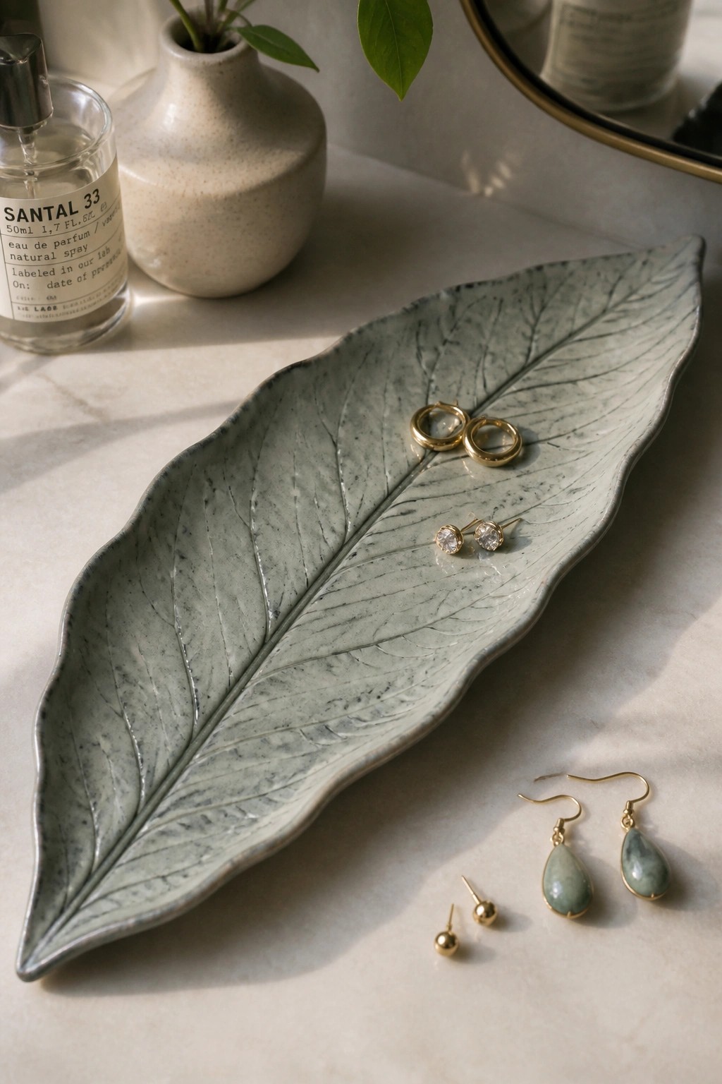 Green leaf-shaped ceramic tray with gold earrings beside perfume bottle on marble