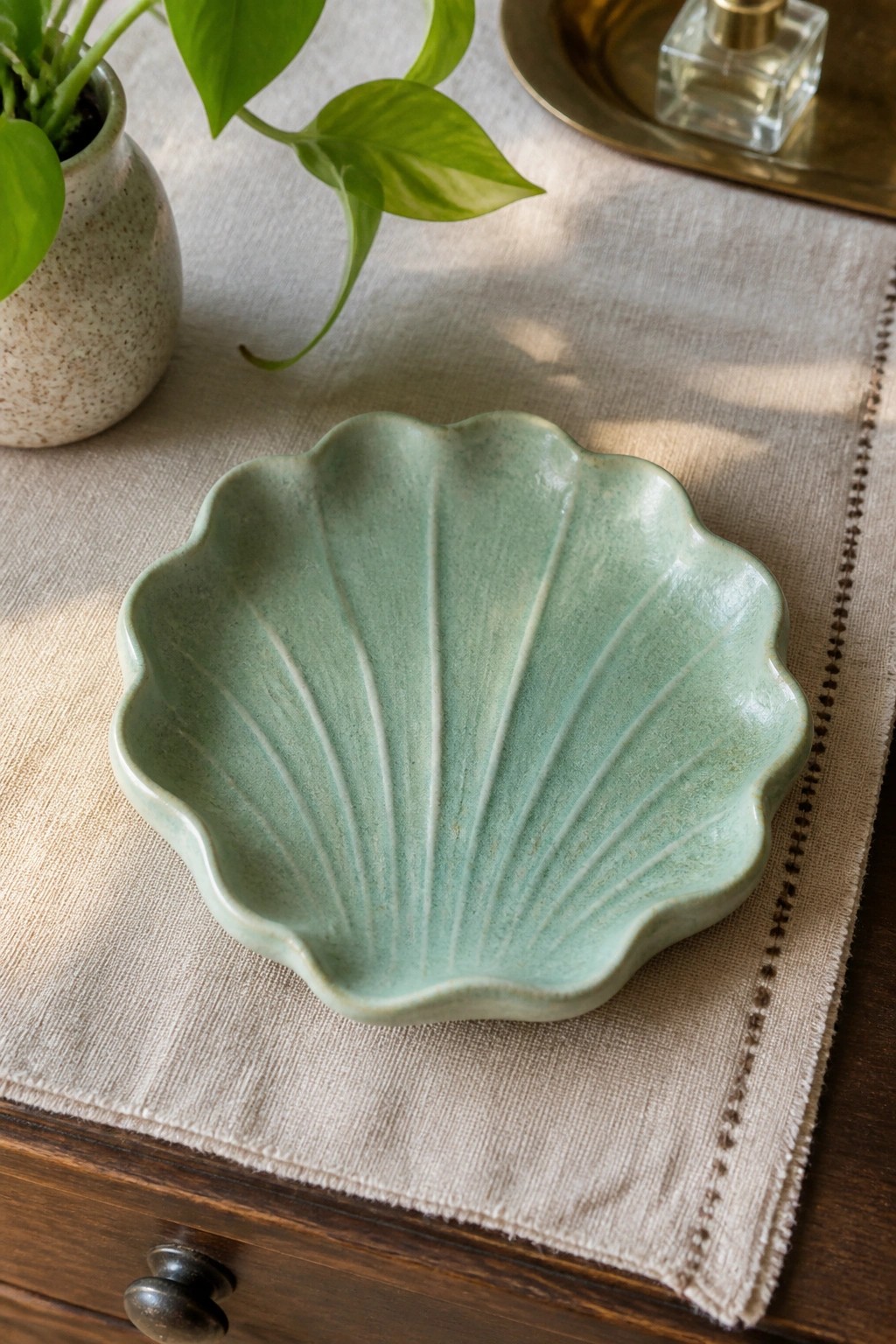 Light green shell-shaped ceramic bowl on beige linen beside potted plant