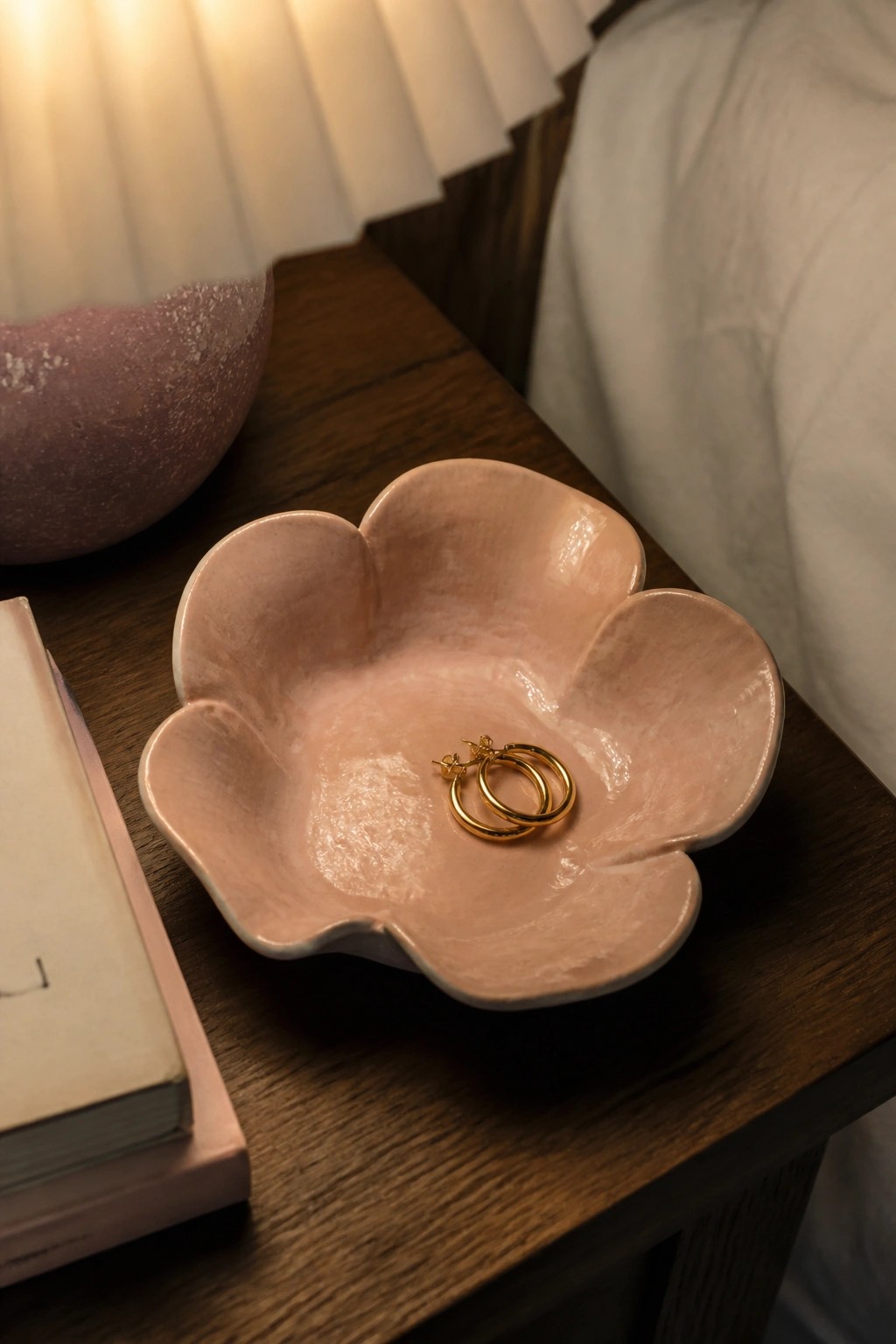 Gold hoop earrings in pink flower-shaped bowl on wooden nightstand beside book and lamp