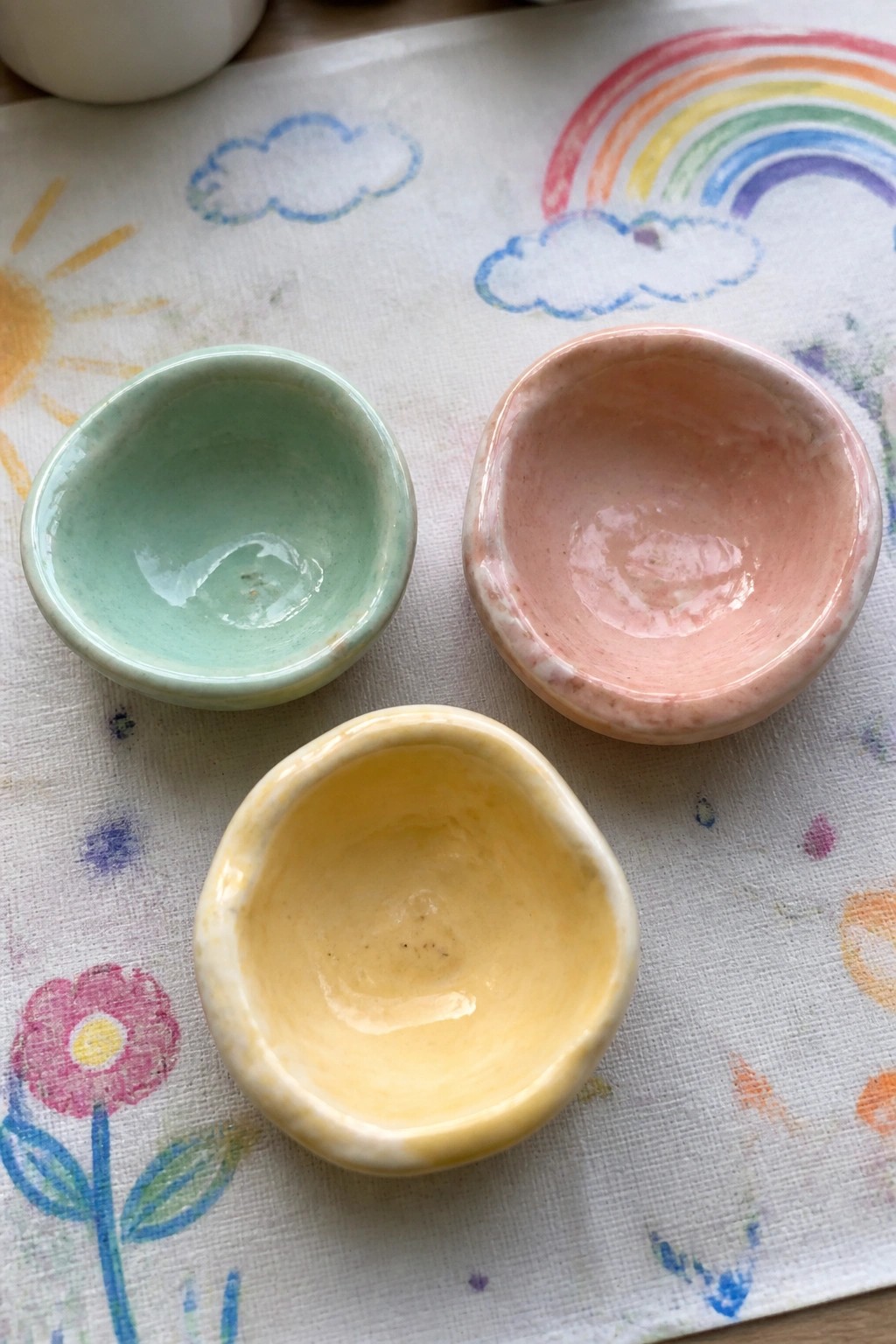 Three small pastel ceramic bowls on colorful rainbow-patterned fabric with sun and flower drawings