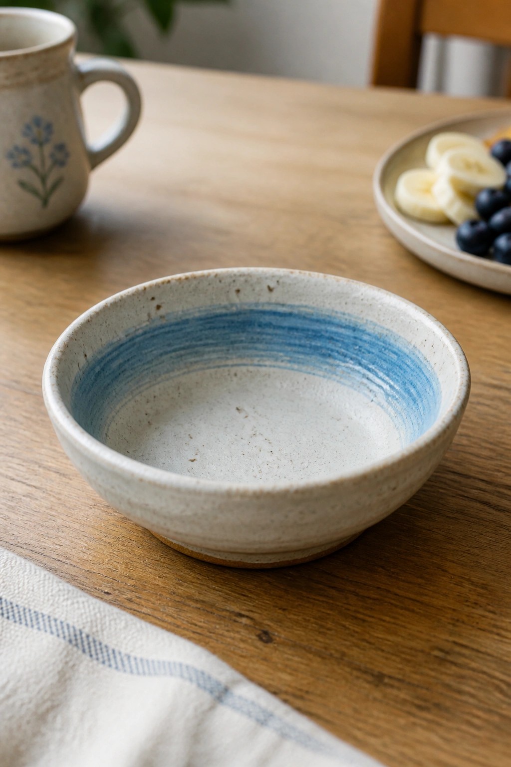 Ceramic bowl with blue stripes beside floral mug and fruit plate on wooden table