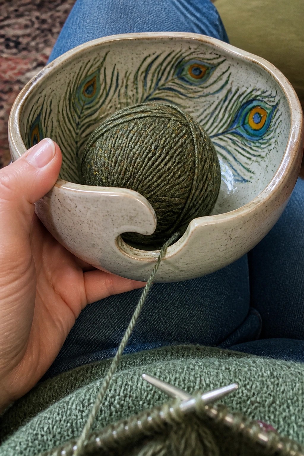 Hand holding peacock-feather ceramic yarn bowl with green yarn and knitting needles.