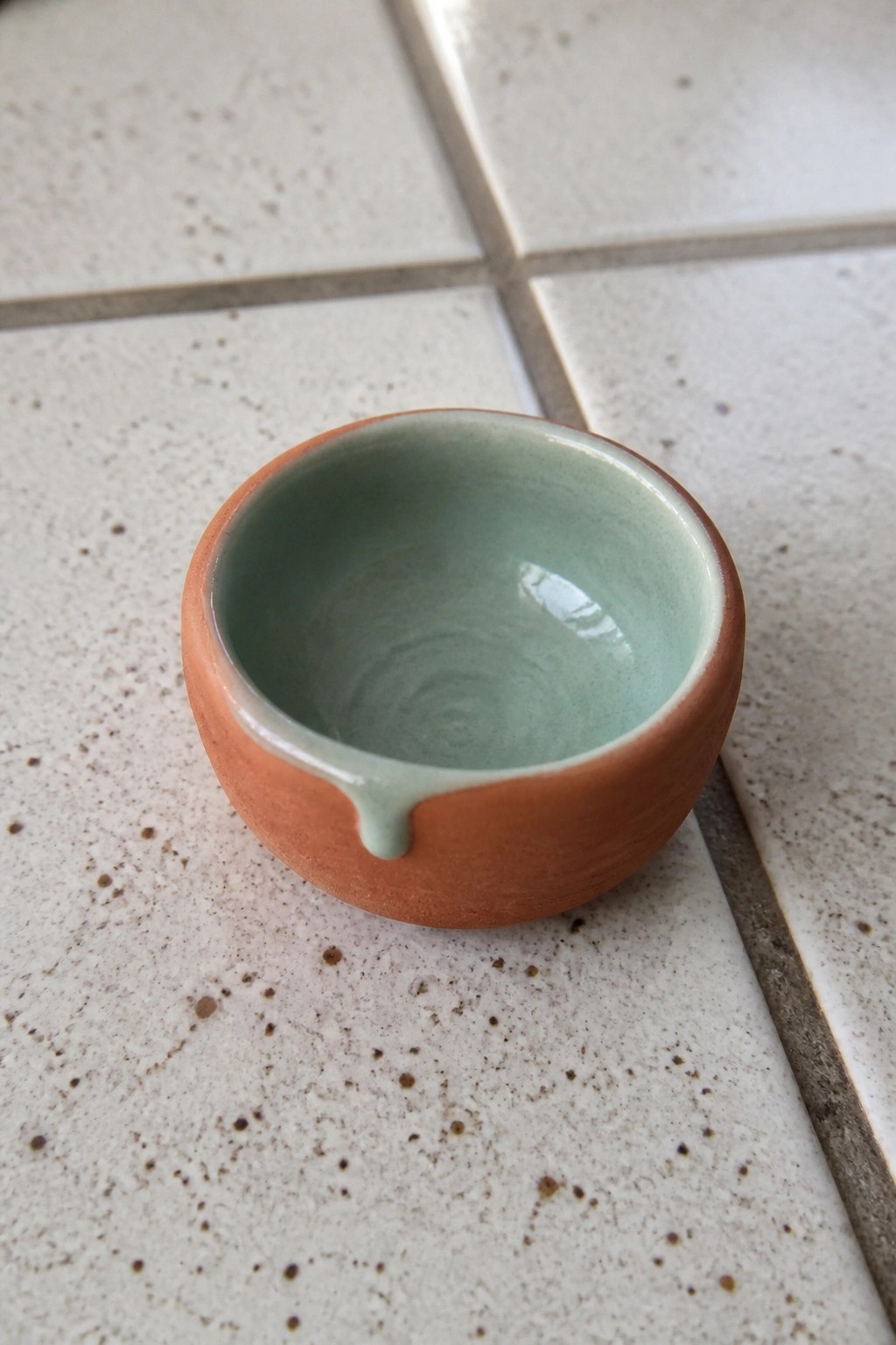 Small terracotta bowl with light green glazed interior and drip on tiles.