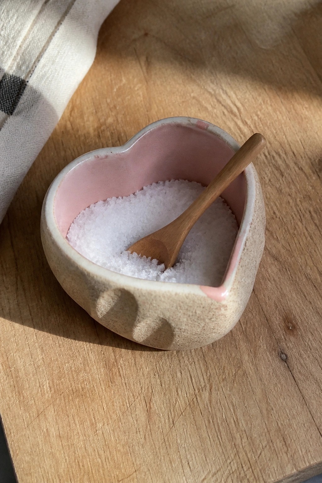 Heart-shaped pink ceramic bowl with white salt and wooden spoon on wooden table