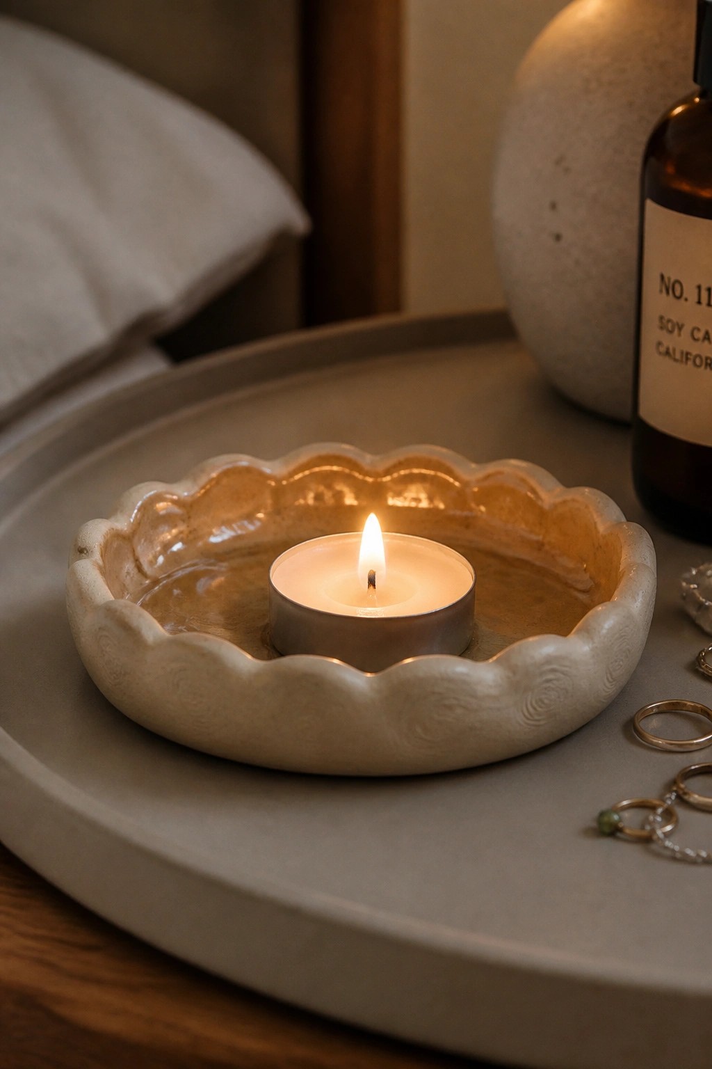 Lit candle in scalloped ceramic bowl on gray tray near jewelry and bottle