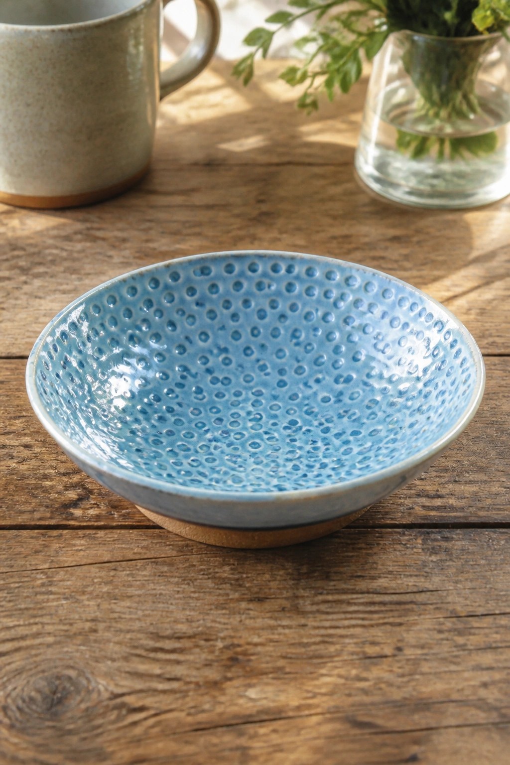 Light blue dimpled ceramic bowl on wooden table with mug and vase