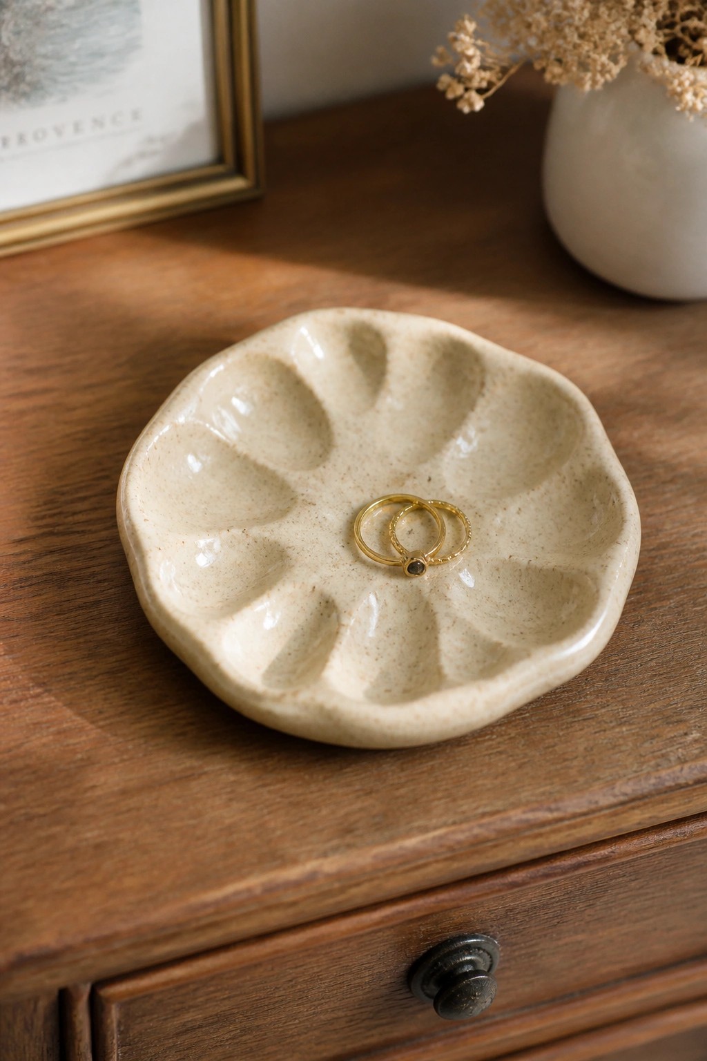Beige scalloped ceramic dish holding two gold rings on wooden dresser.