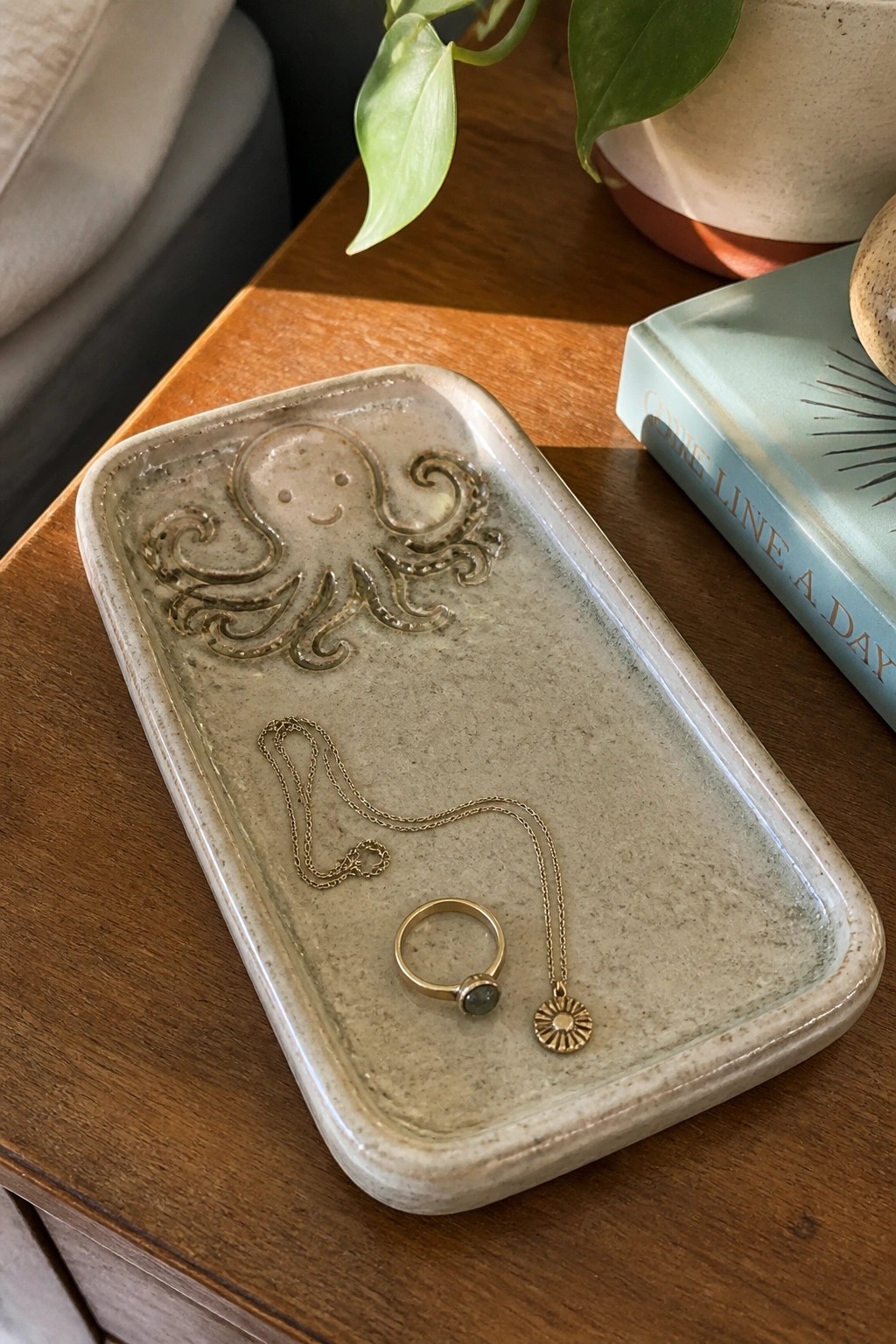 Rectangular ceramic tray with raised octopus design and smiling face holding gold jewelry on a wooden surface.