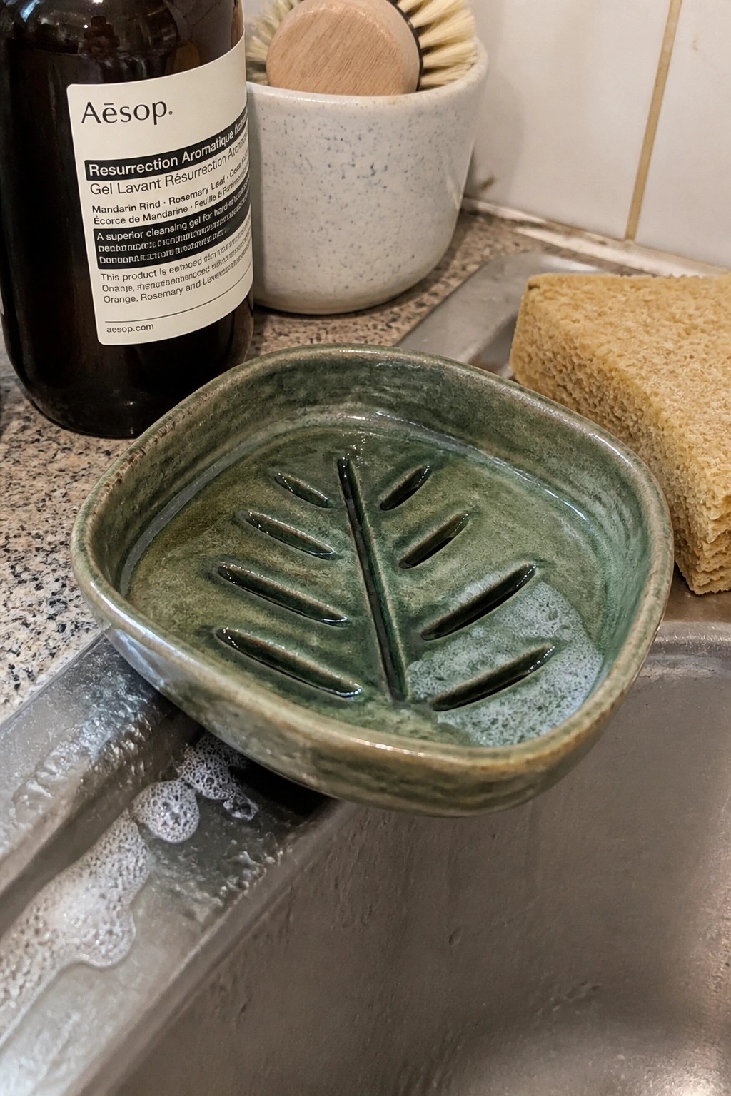 Green ceramic leaf soap dish on sink edge with Aesop bottle and sponge nearby
