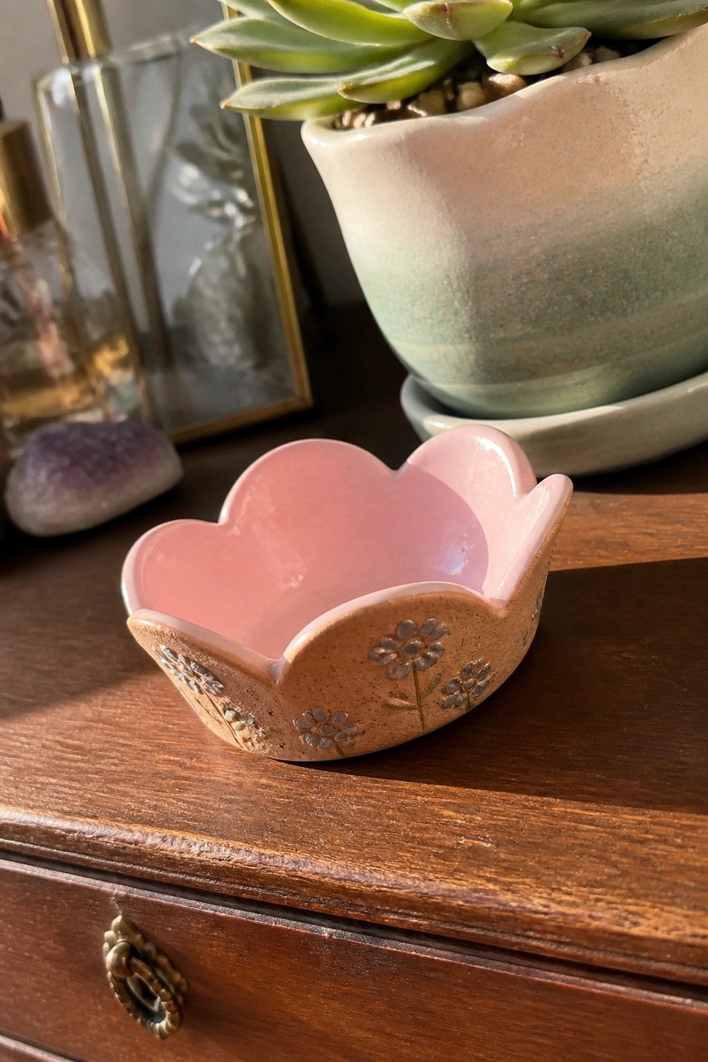 A small pink ceramic bowl with scalloped petal edges sits on a wooden surface, featuring painted silver flowers on its brown exterior.