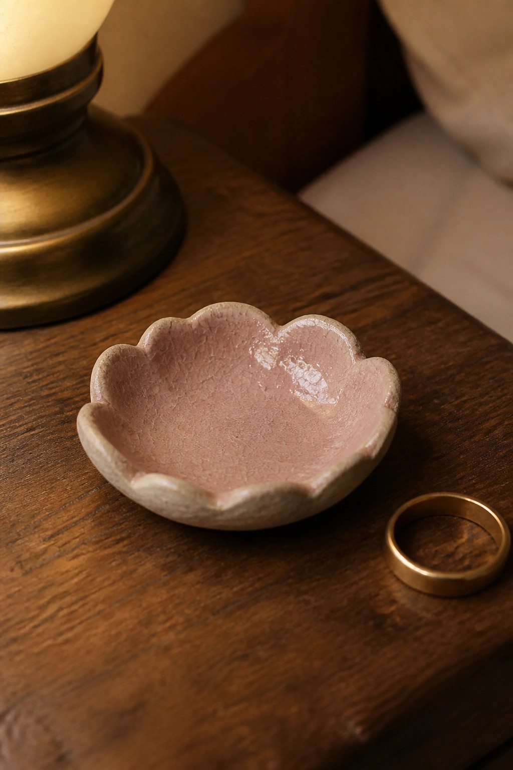 Crackled pink ceramic bowl and gold ring on dark wooden table near lamp.