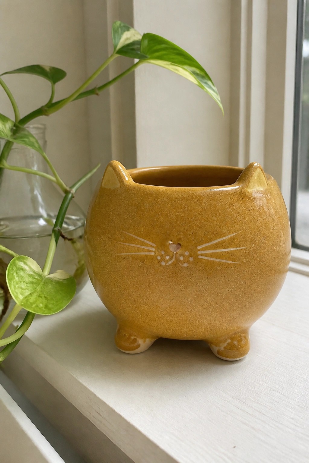 Yellow cat-shaped ceramic planter with etched face on white windowsill beside green plant