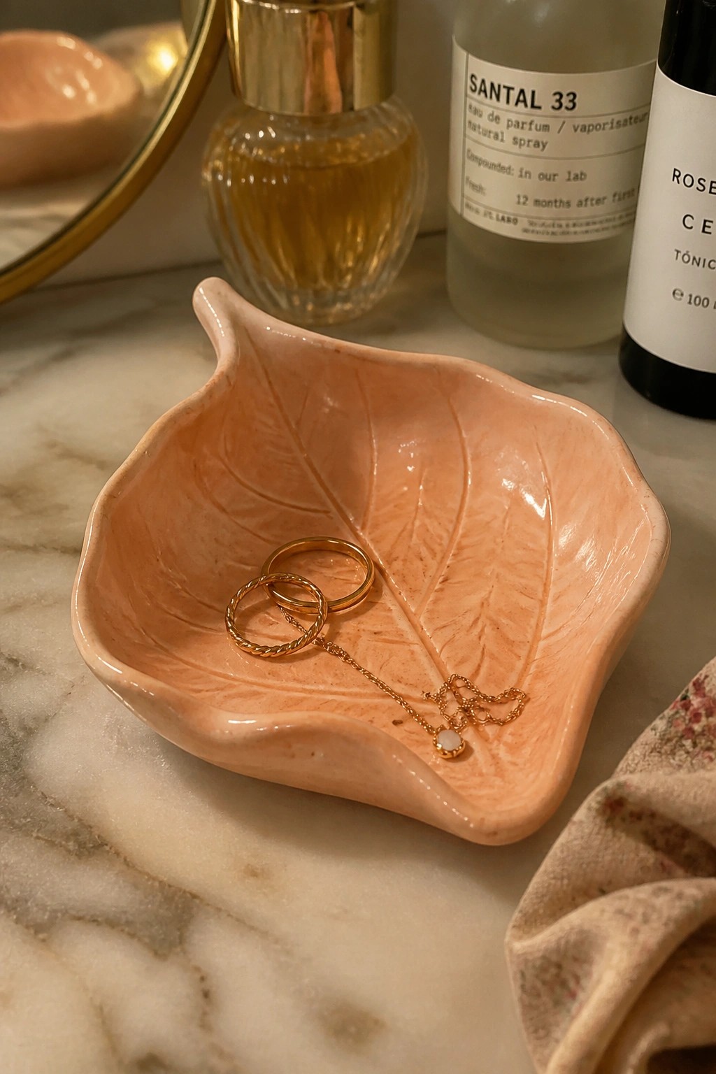 Pink leaf-shaped ceramic dish holding gold rings and pendant necklace on marble.