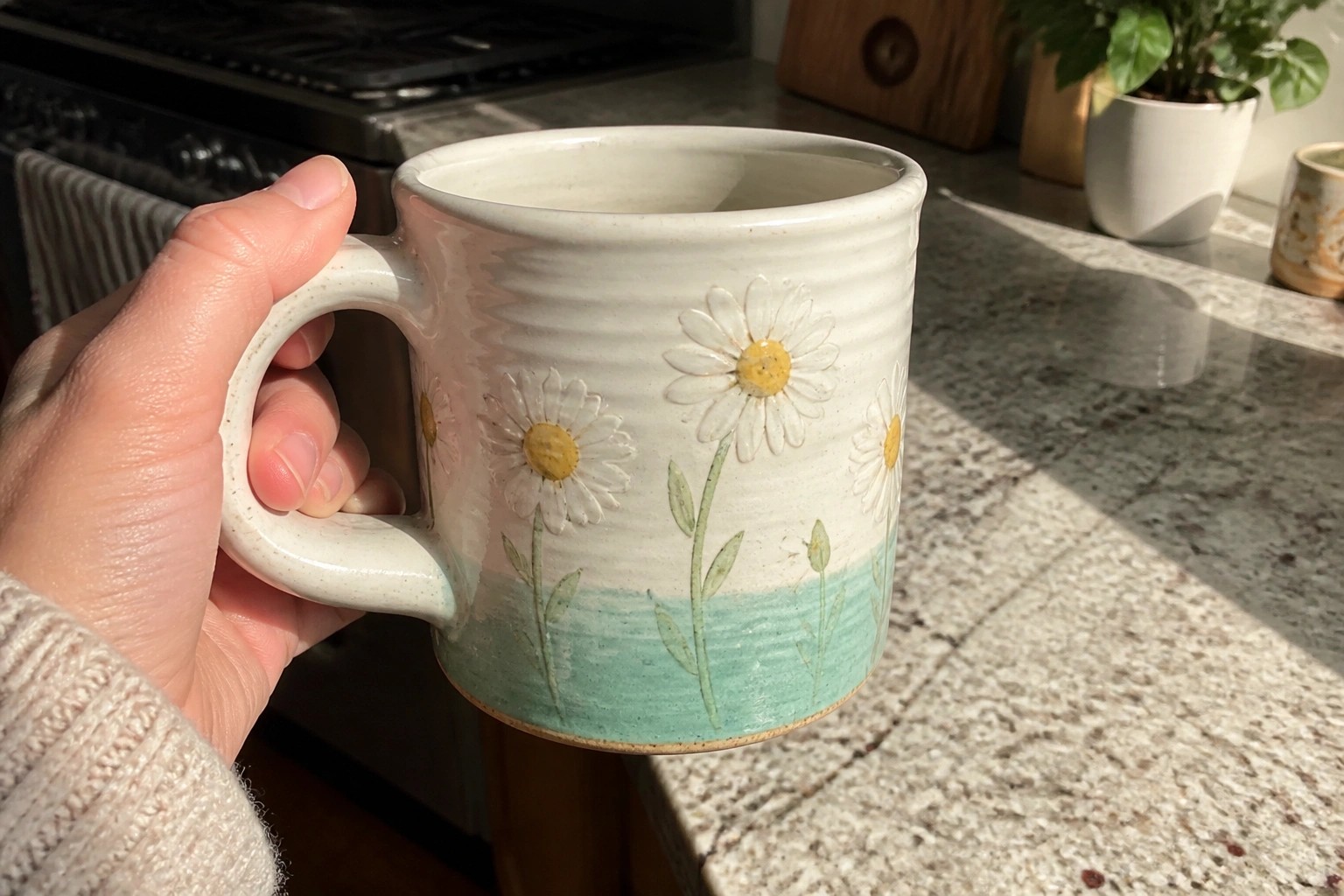 A hand holds a white ceramic mug with three painted daisies and a turquoise glaze on the lower half.