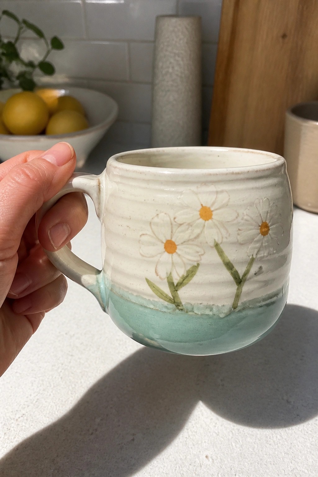 A hand holds a white ceramic mug with three painted daisies and a turquoise glaze on the lower half.