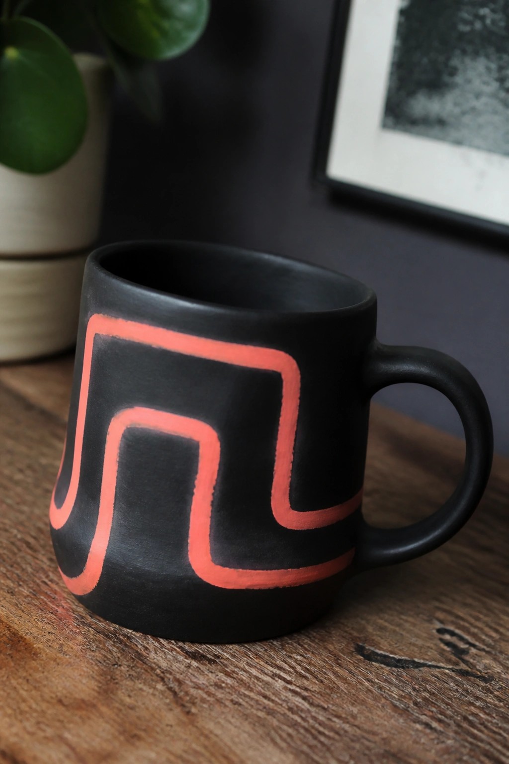 Black mug with red geometric design on wooden table, plant in background