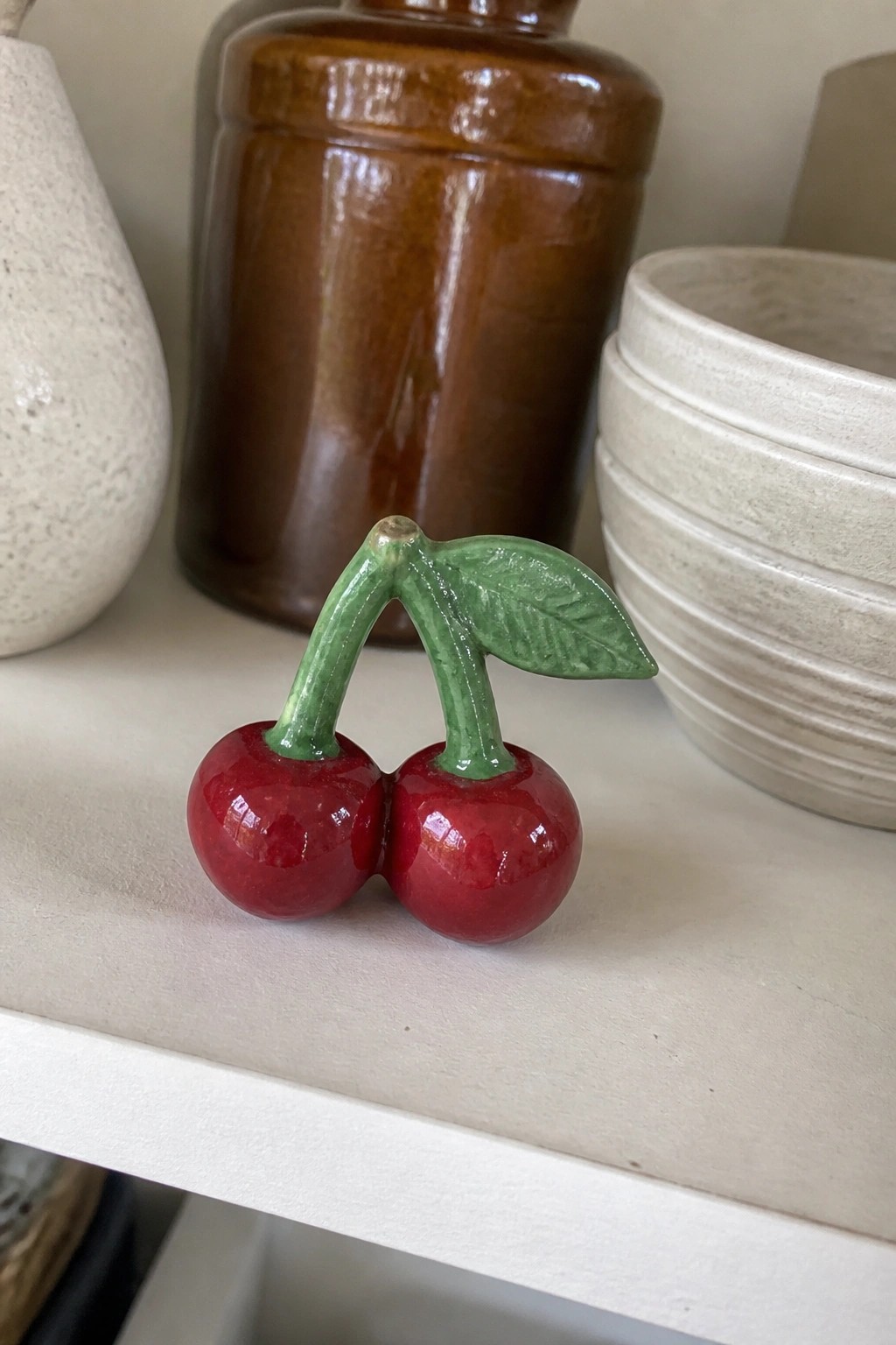Two glossy red ceramic cherries with green stems and leaf on white shelf