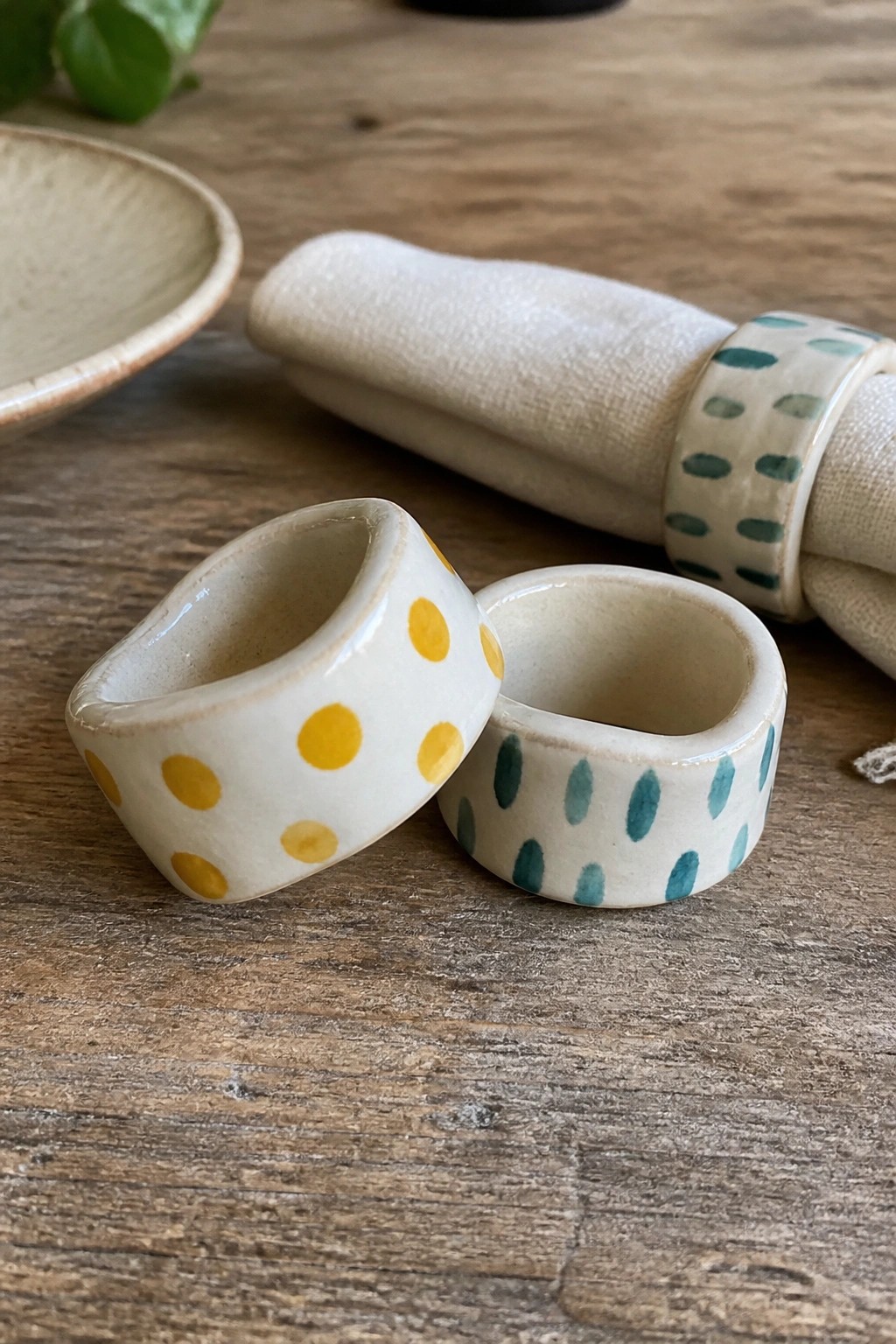 Two ceramic napkin rings with yellow and teal dots beside rolled cloth on wood