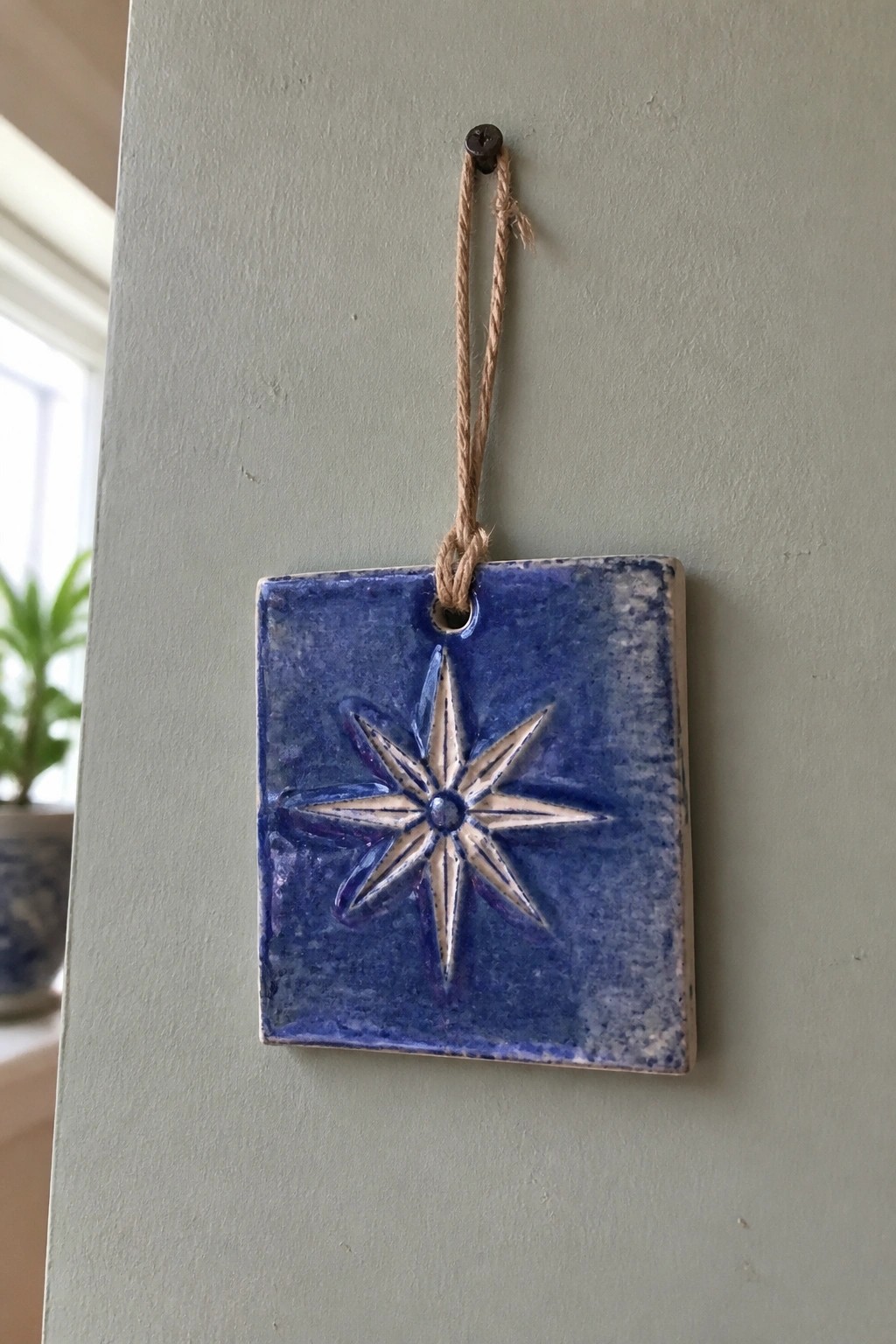 Blue square tile with raised white star hanging from twine on green wall.