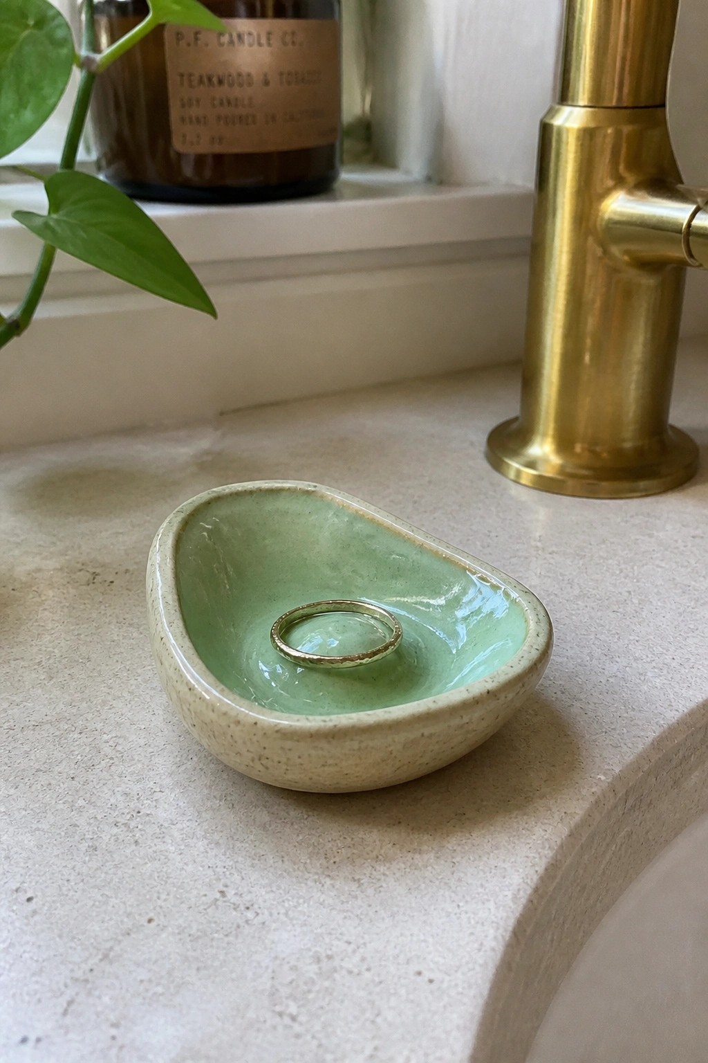 Small green ceramic dish holding gold ring on beige countertop near brass faucet