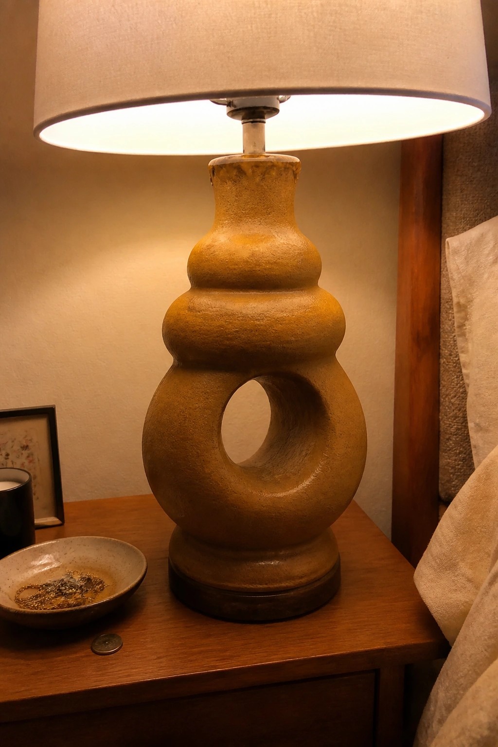 Lit yellow ceramic lamp with circular opening on wooden bedside table with jewelry bowl