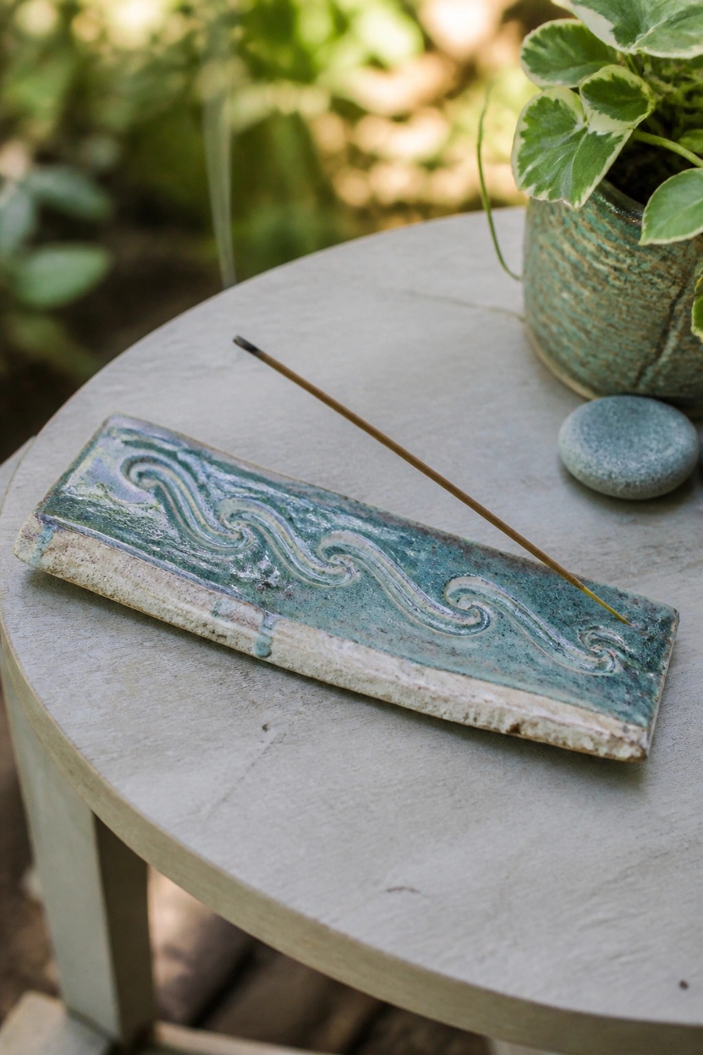 Ceramic wave-patterned incense holder with stick on round table beside potted plant.