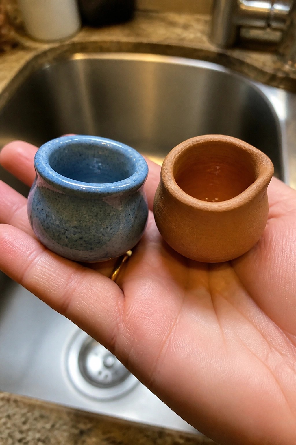 Hand holding blue speckled ceramic cup and brown terracotta pot over sink.
