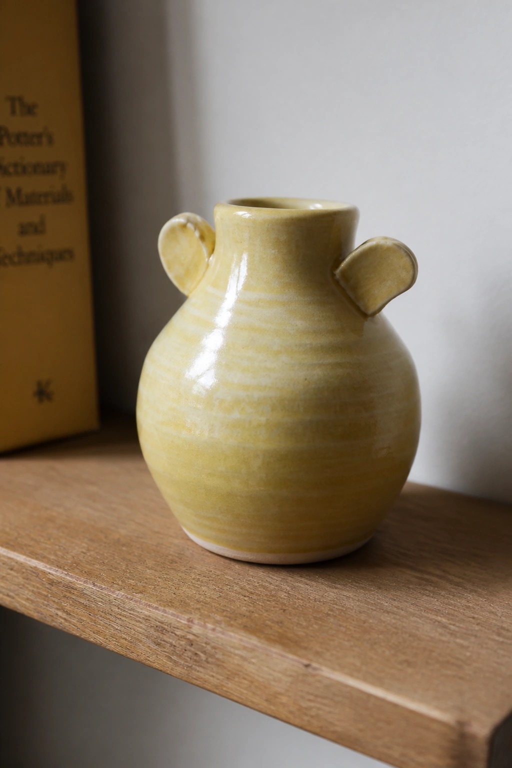 Yellow ceramic vase with two handles on wooden shelf beside book