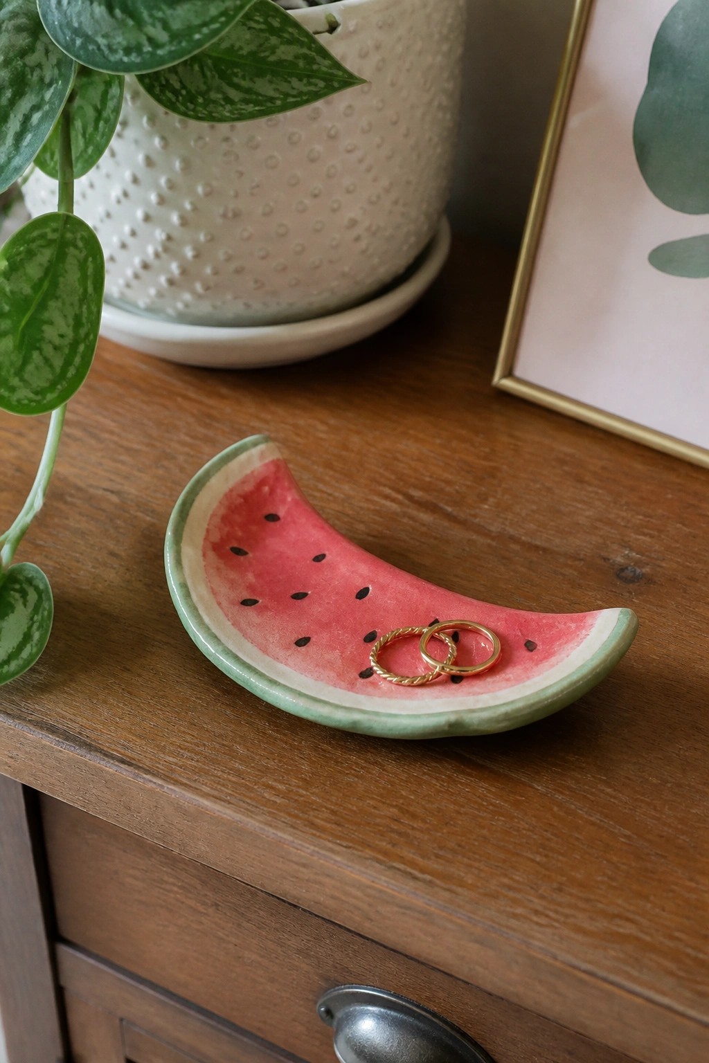 A small ceramic dish shaped like a watermelon slice holds two gold rings on a wooden surface.