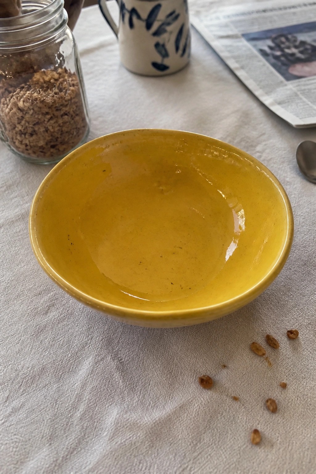 Yellow bowl on white tablecloth with granola jar, blue mug, newspaper, and scattered oats.