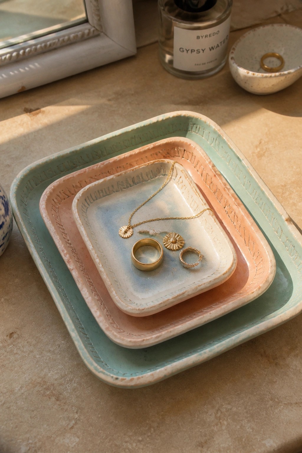 Nested pastel ceramic trays with gold jewelry beside Byredo perfume bottle