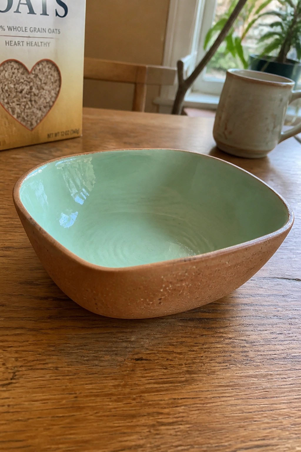 Green ceramic bowl on wooden table beside oats box and ceramic mug