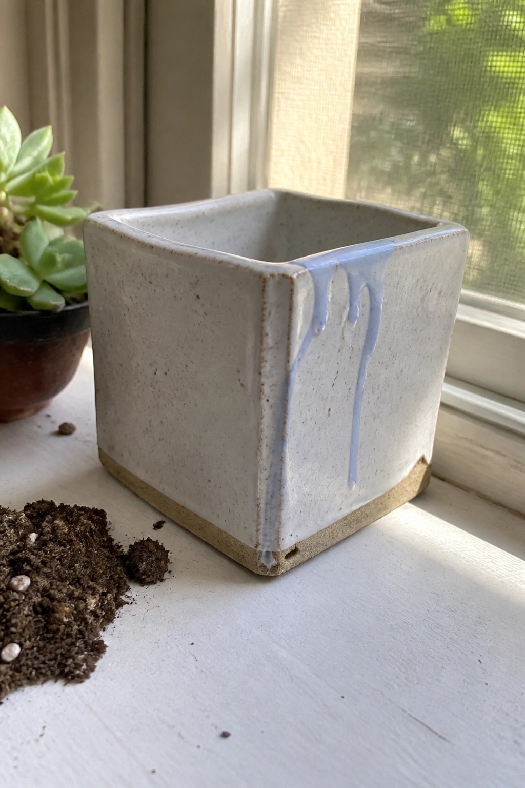 Square white ceramic pot with blue glaze drips on windowsill beside succulent and soil