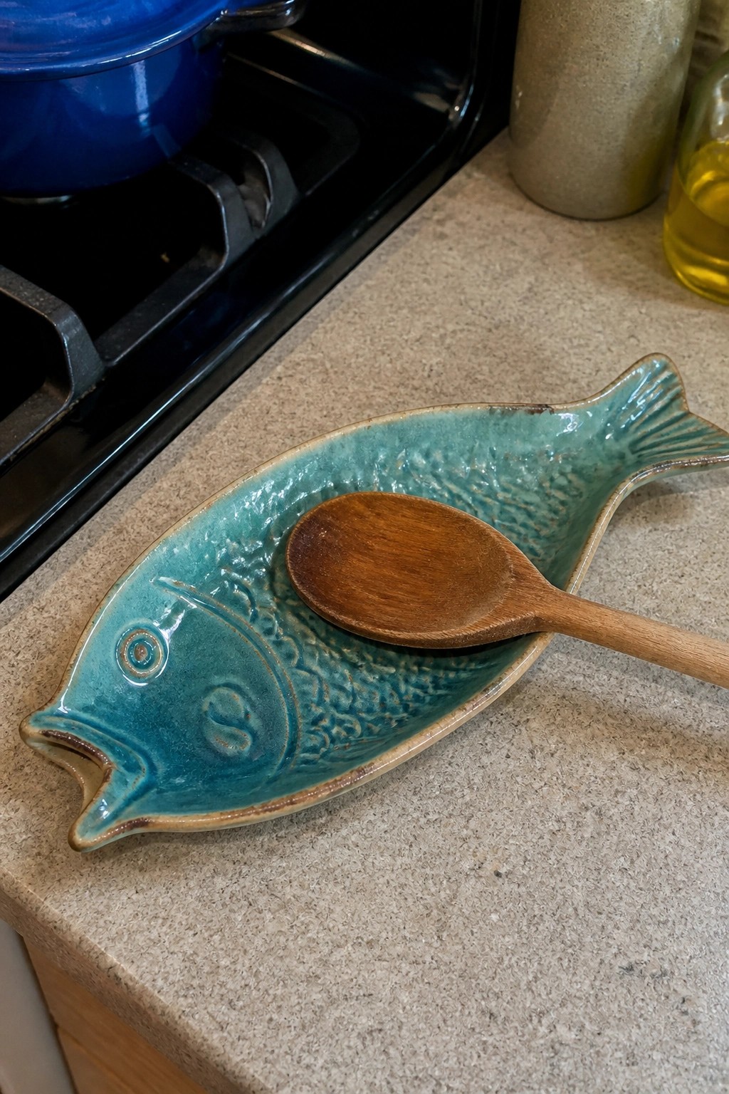 Turquoise fish-shaped ceramic dish holding wooden spoon on kitchen countertop near stove