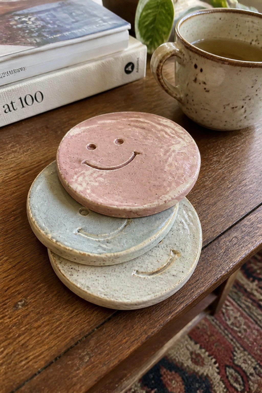 Three smiley face ceramic coasters stacked on wooden table beside speckled mug and books.