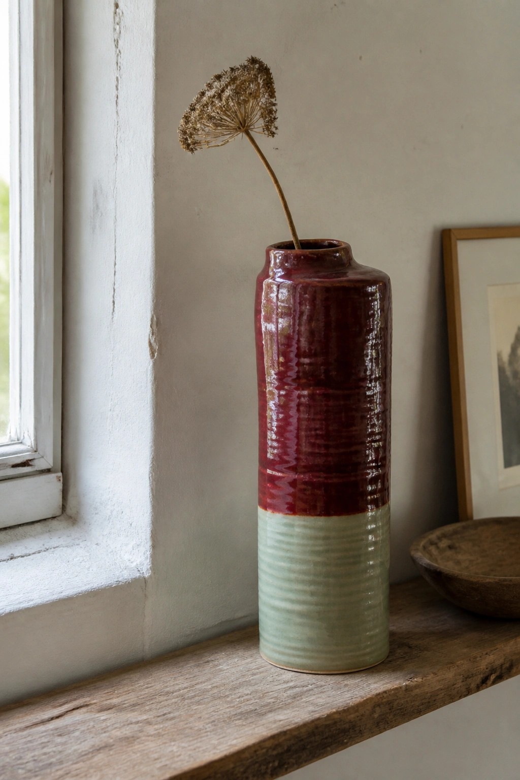 Red and green ribbed ceramic vase with dried flower on wooden windowsill