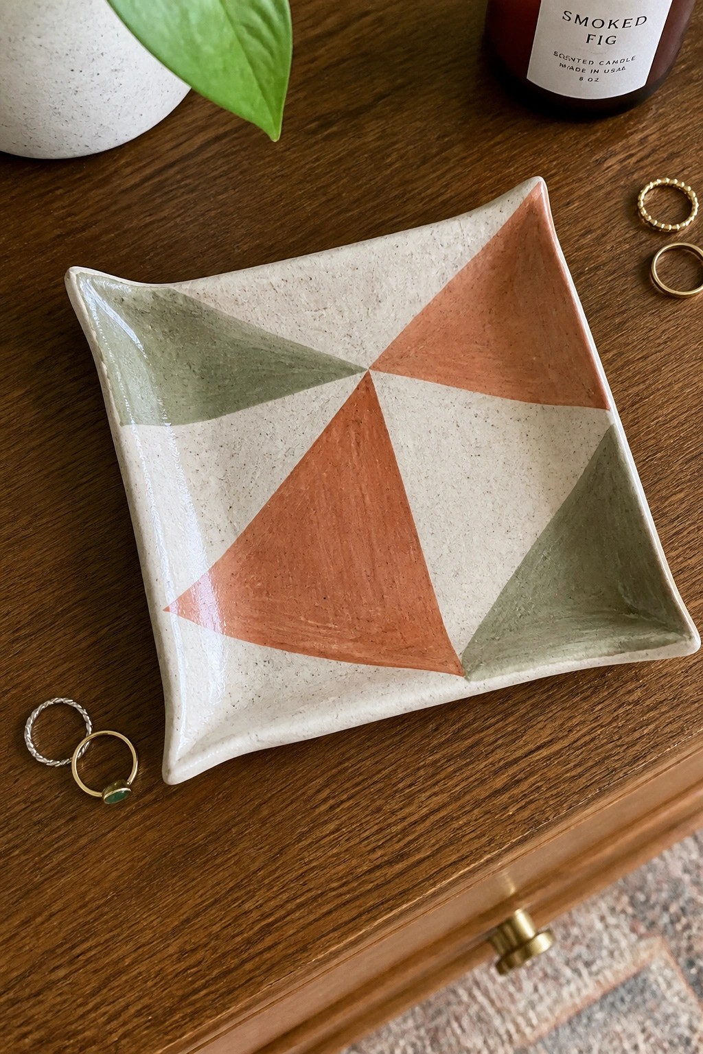 A small ceramic tray with orange, cream, and green triangular glaze sections rests on a wooden surface next to three rings.