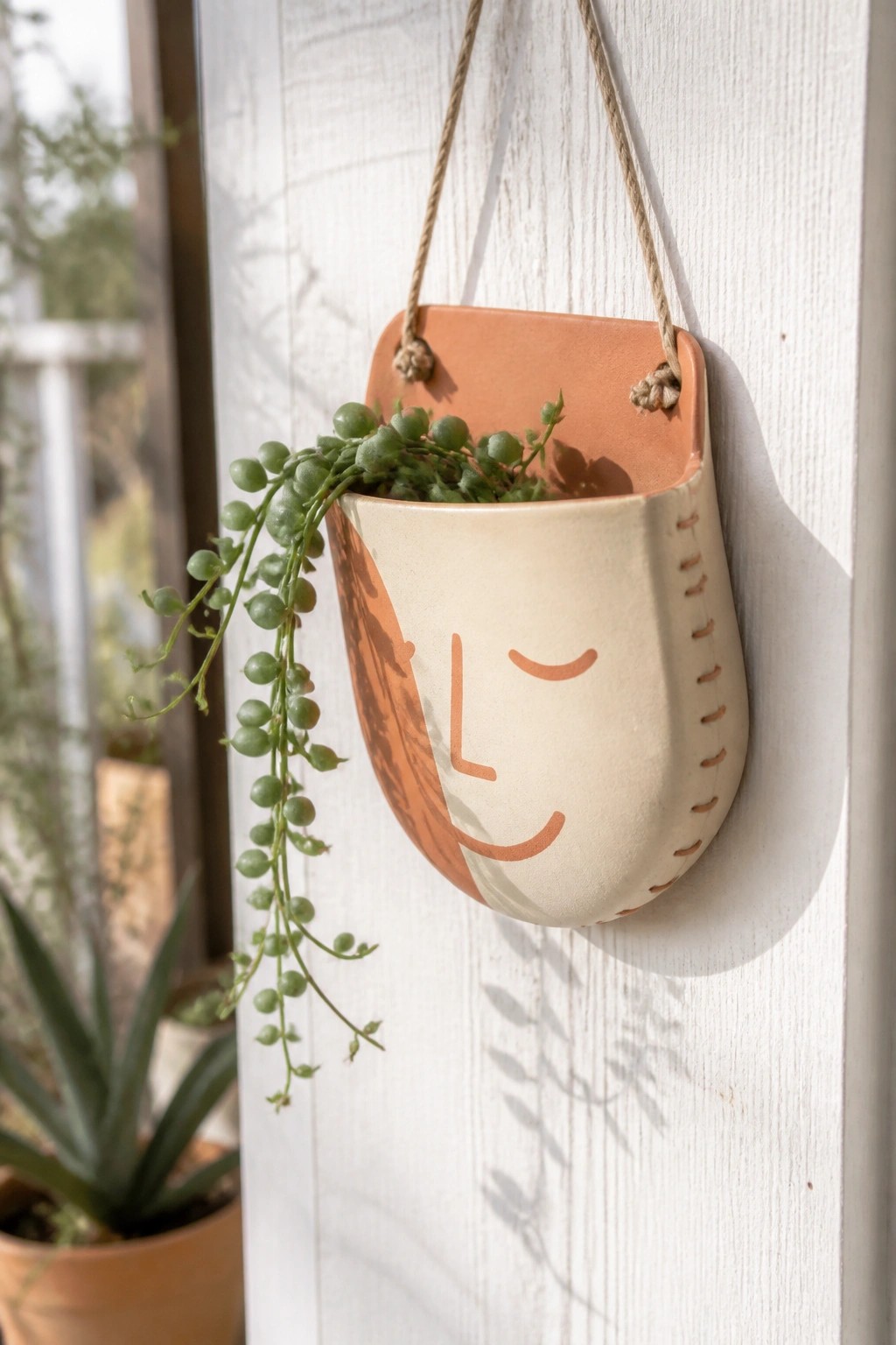 Hanging two-toned ceramic face planter with trailing green succulent on white wall.