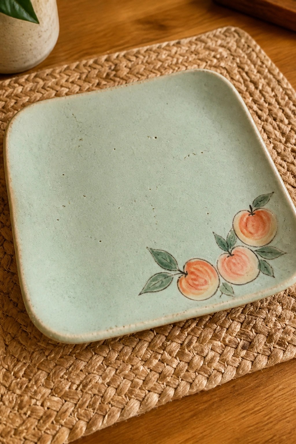 Mint green square ceramic plate with painted peaches on woven placemat atop wooden table