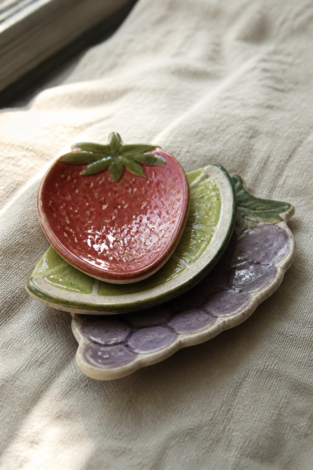 Stacked ceramic fruit dishes: red strawberry atop green lime over purple grapes on linen.