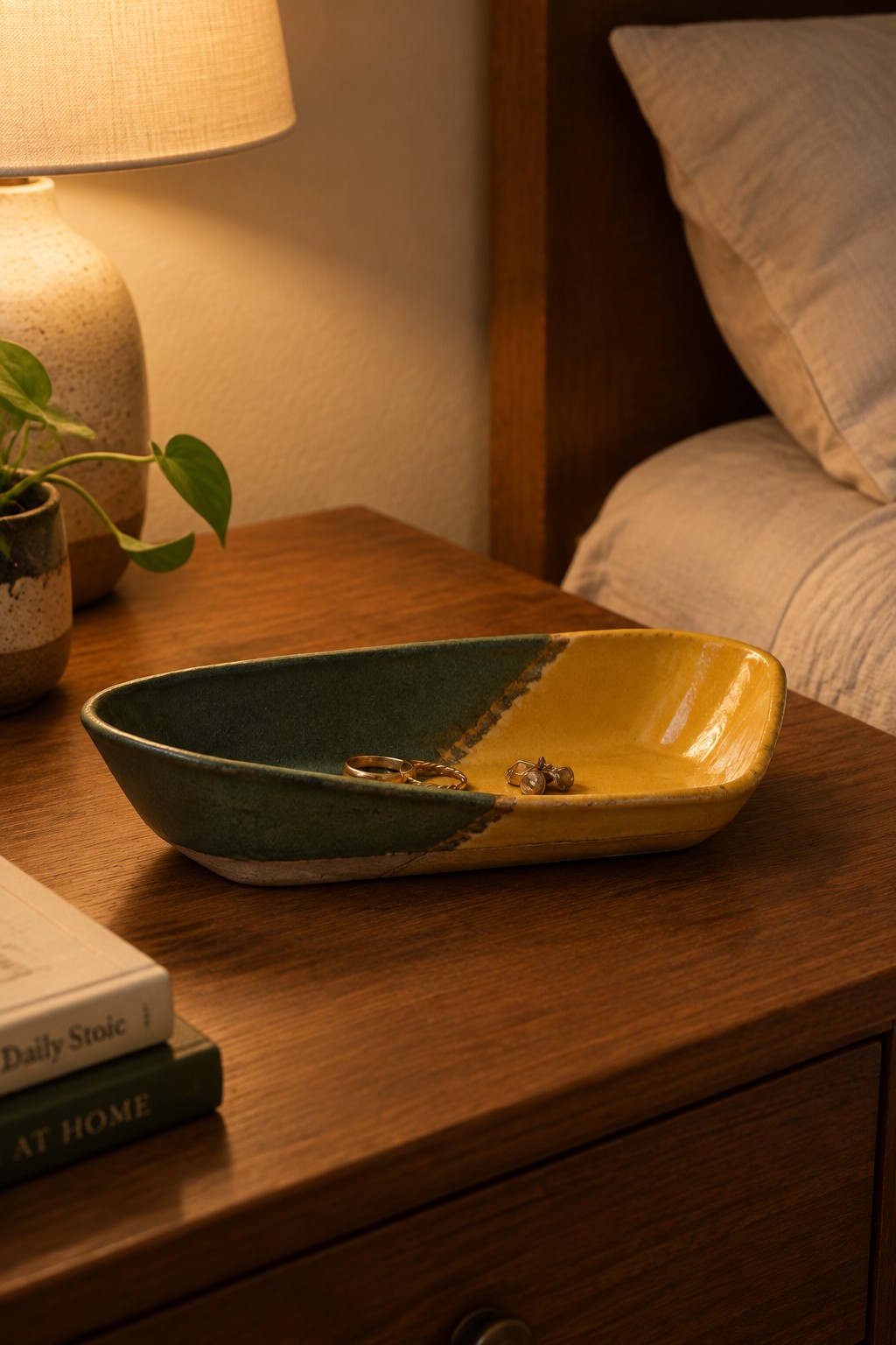 Two-toned green-yellow ceramic bowl with jewelry on wooden nightstand beside lamp and books