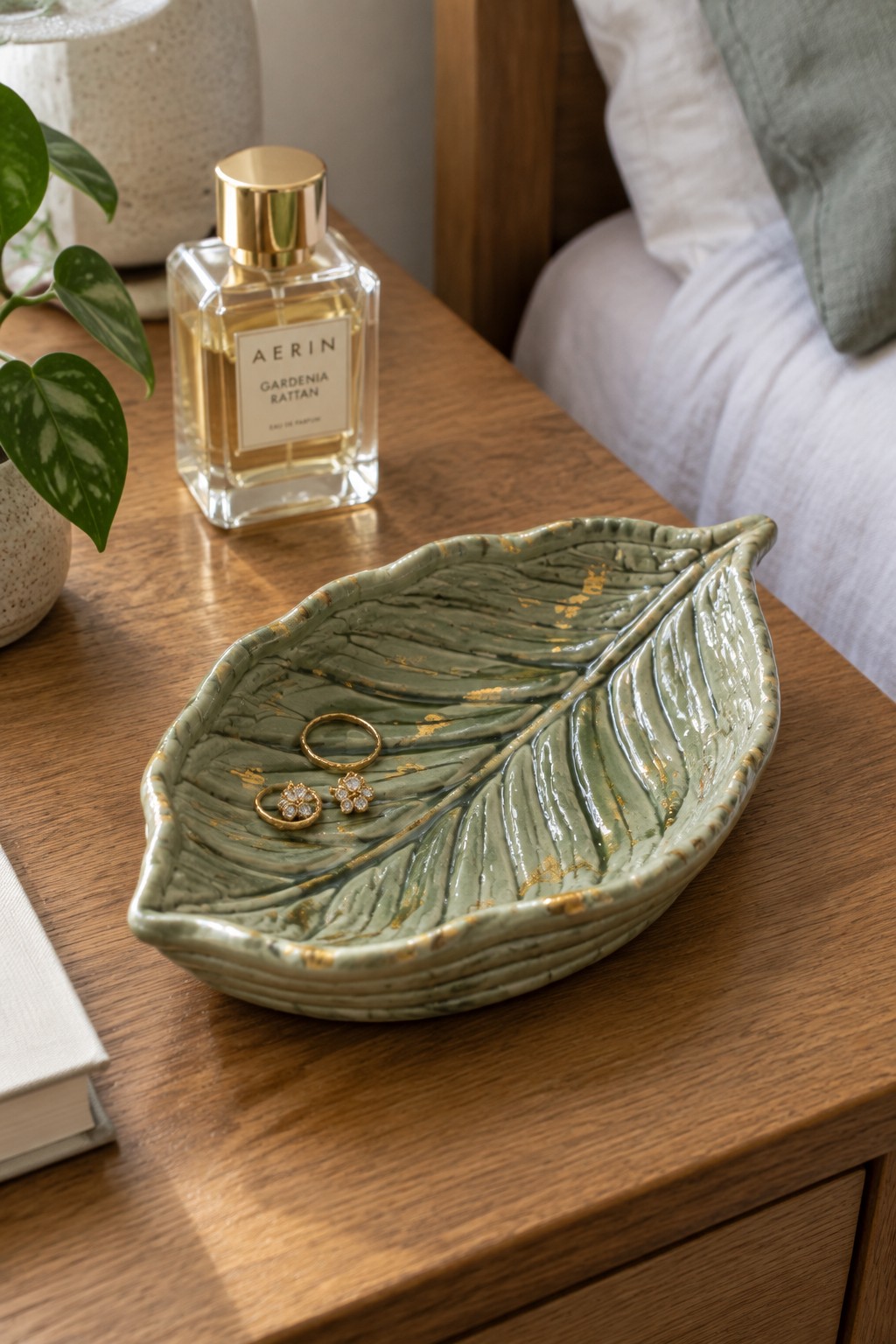 Green ceramic leaf tray with gold jewelry beside perfume bottle on wooden nightstand