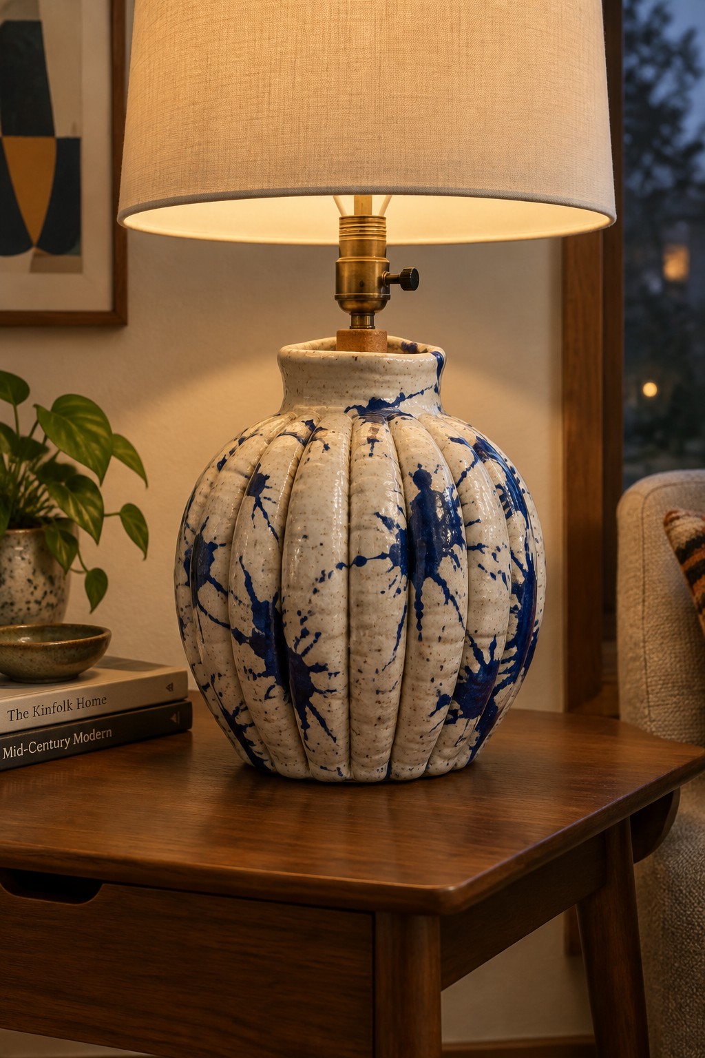 Blue-splattered ribbed ceramic lamp glowing on wooden table beside books and plant