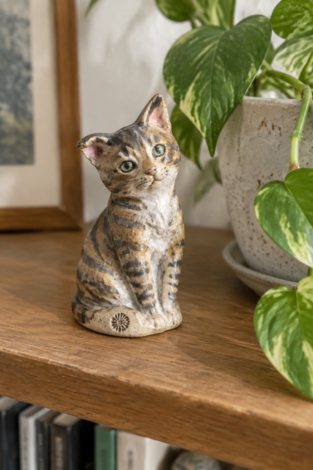 Ceramic tabby cat figurine on wooden shelf beside variegated potted plant
