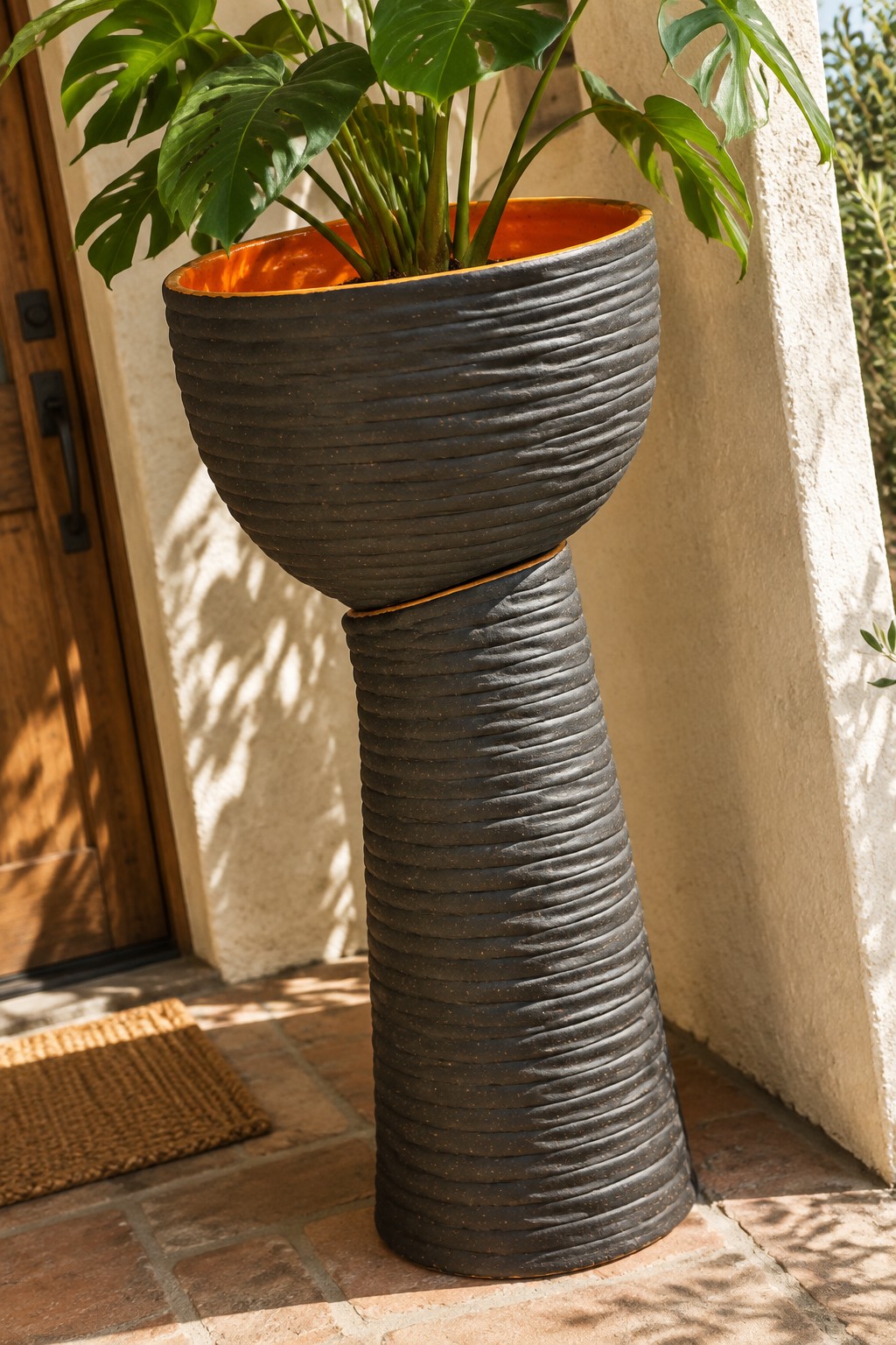Monstera plant in tall ribbed dark planter with orange interior on brick patio