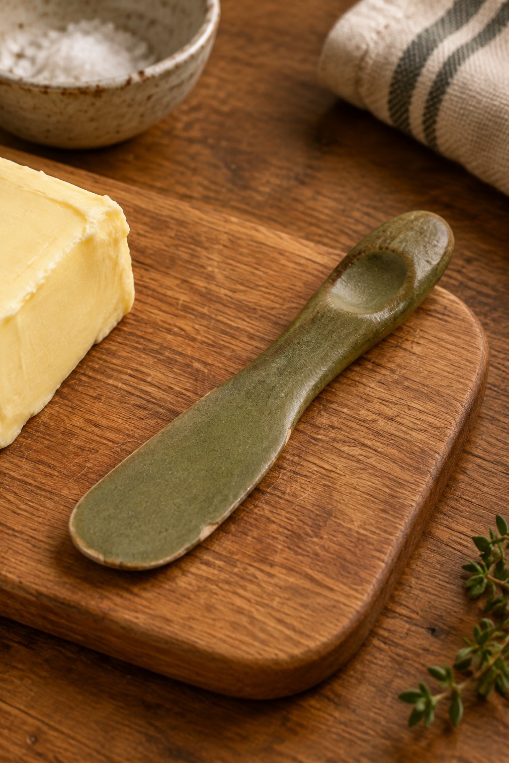 Yellow butter block with green ceramic knife on wooden board beside salt bowl and thyme
