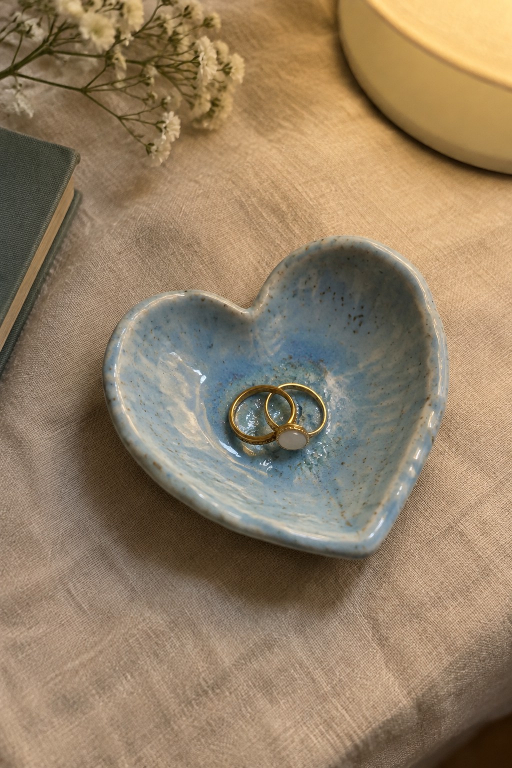 Blue heart-shaped ceramic bowl holding two gold rings on beige fabric